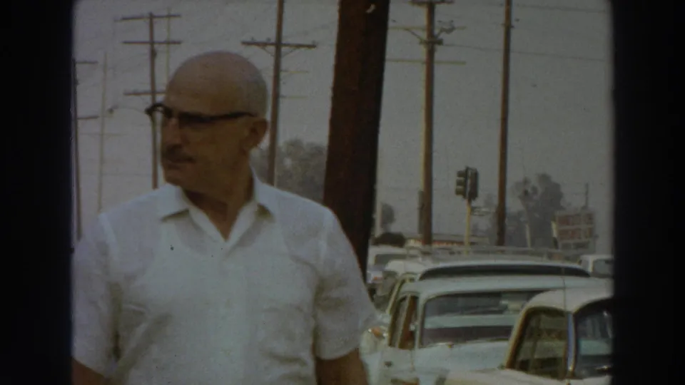 The man glances to the side while passing the camera on a 1966 Puerto Rico sidewalk lined with parked cars.