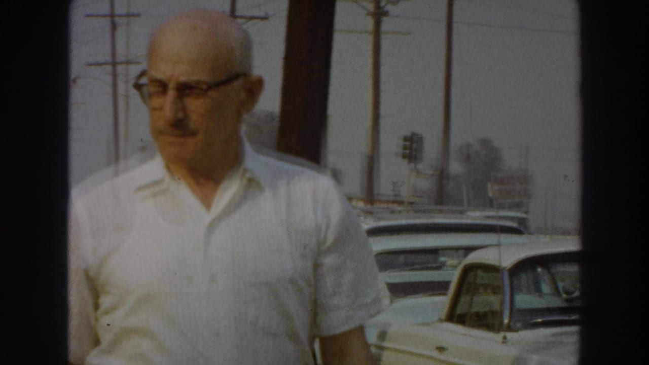 Man in a white short-sleeve shirt walks near parked mid-century cars and utility poles.