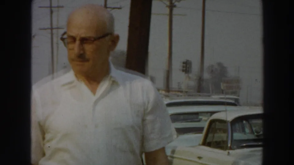 In Puerto Rico, 1966, a man wearing glasses and a white shirt walks along a sunlit roadside with cars and utility poles behind him.