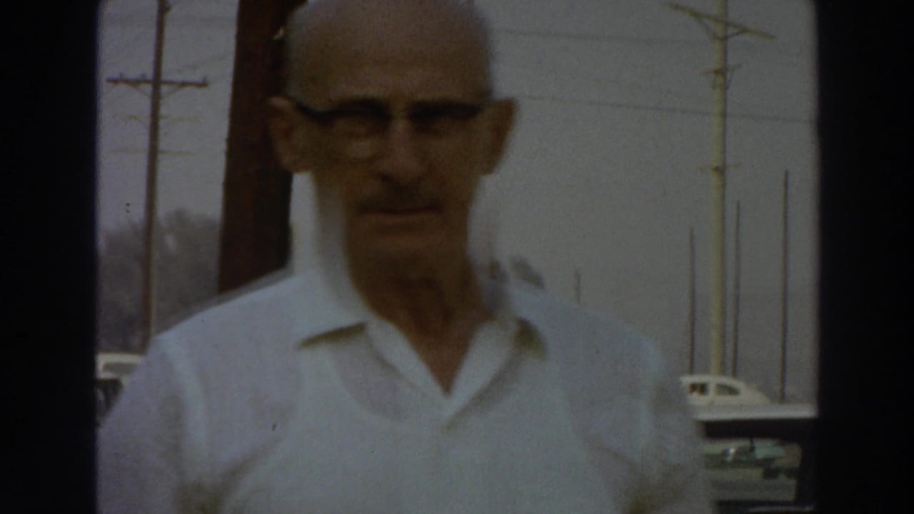 The man in glasses approaches the camera in a 1966 Puerto Rico street scene, with parked cars and roadside poles in the background.