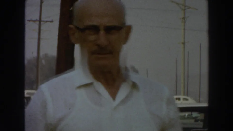 The man in glasses approaches the camera in a 1966 Puerto Rico street scene, with parked cars and roadside poles in the background.