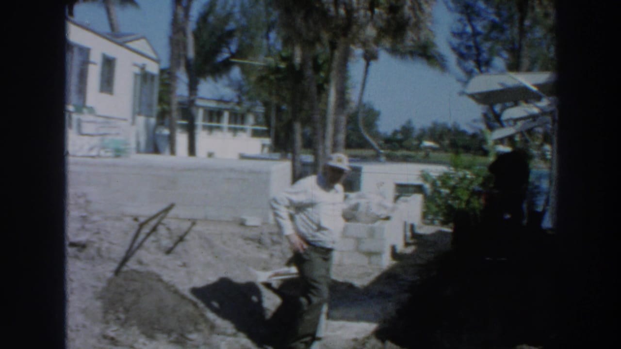 In a sunlit Puerto Rico beachside or outdoor setting, the man stands near a low wall by a white building and palm trees.