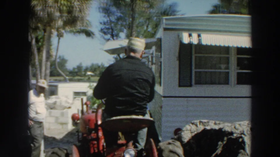 1966 Puerto Rico home-movie frame showing a man from behind on a red tractor beside a white trailer and another worker nearby.