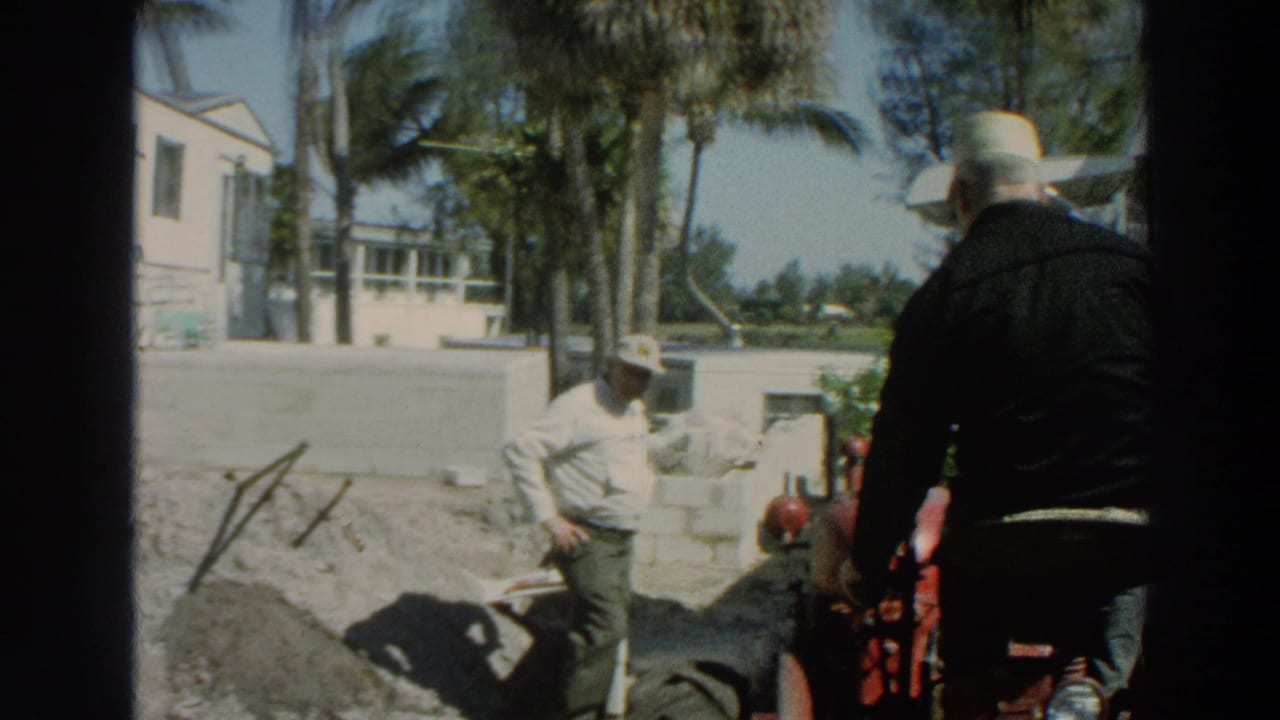 1966 Puerto Rico frame showing two men near a red tractor, dirt piles, cinder blocks, palm trees, and a white trailer at a work site.
