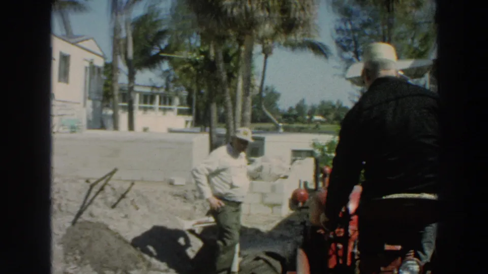 1966 Puerto Rico frame showing two men near a red tractor, dirt piles, cinder blocks, palm trees, and a white trailer at a work site.