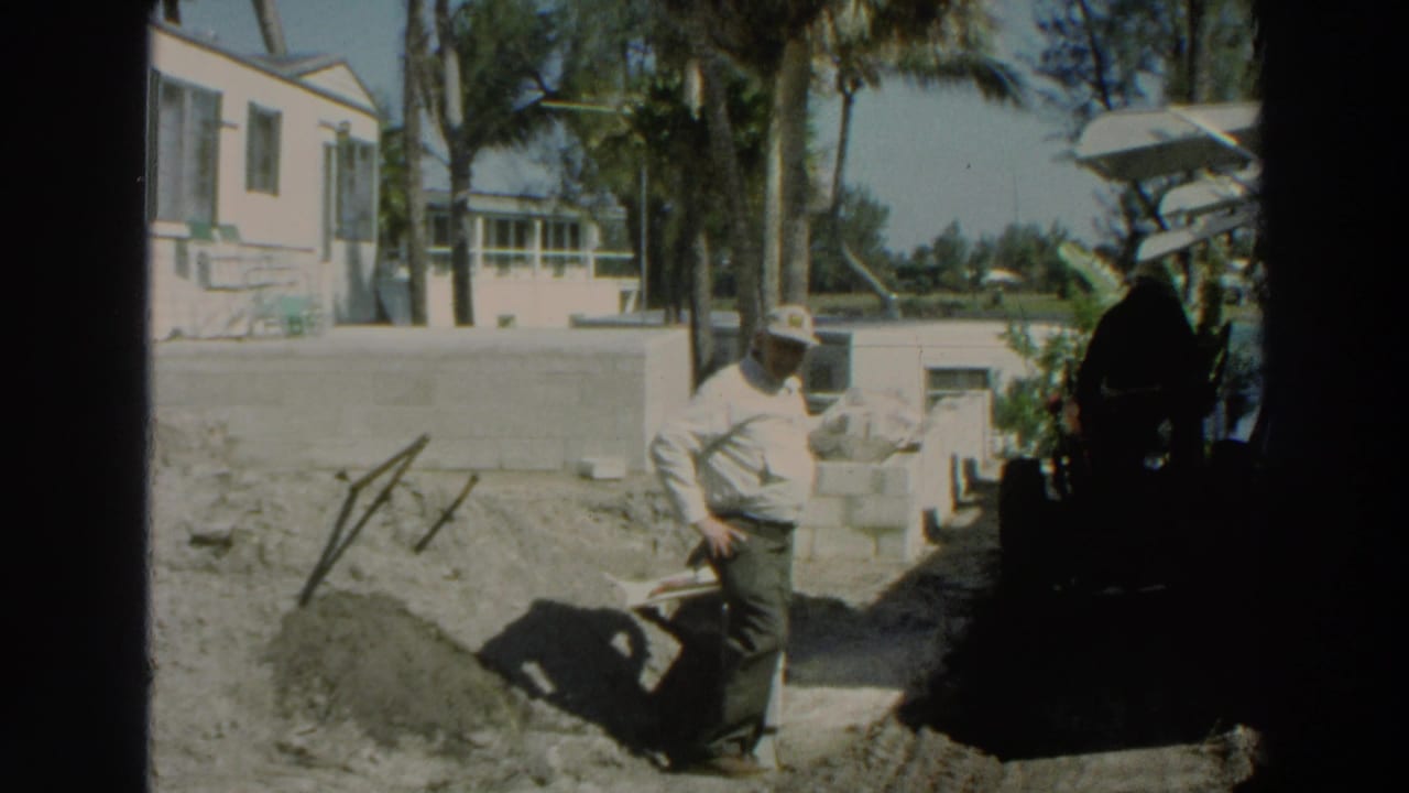 Man in a hat stands with a shovel near a concrete block wall and soil pile.