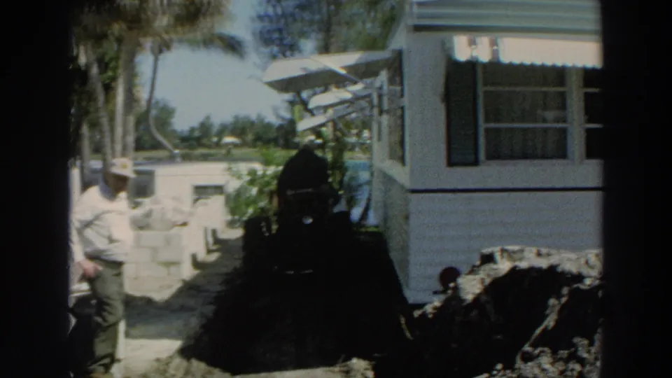 Puerto Rico 1966 8mm frame showing the man partly at left beside masonry work, soil piles, a residence, and tropical trees.