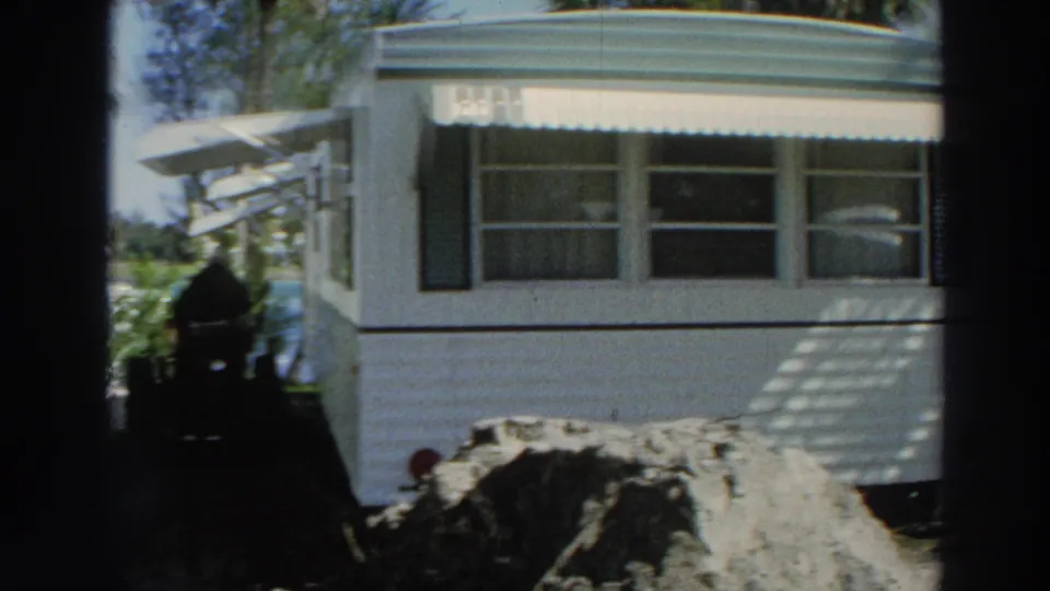 1966 Puerto Rico home-movie frame centered on a residence, striped awning, soil piles, and shadowed construction equipment.