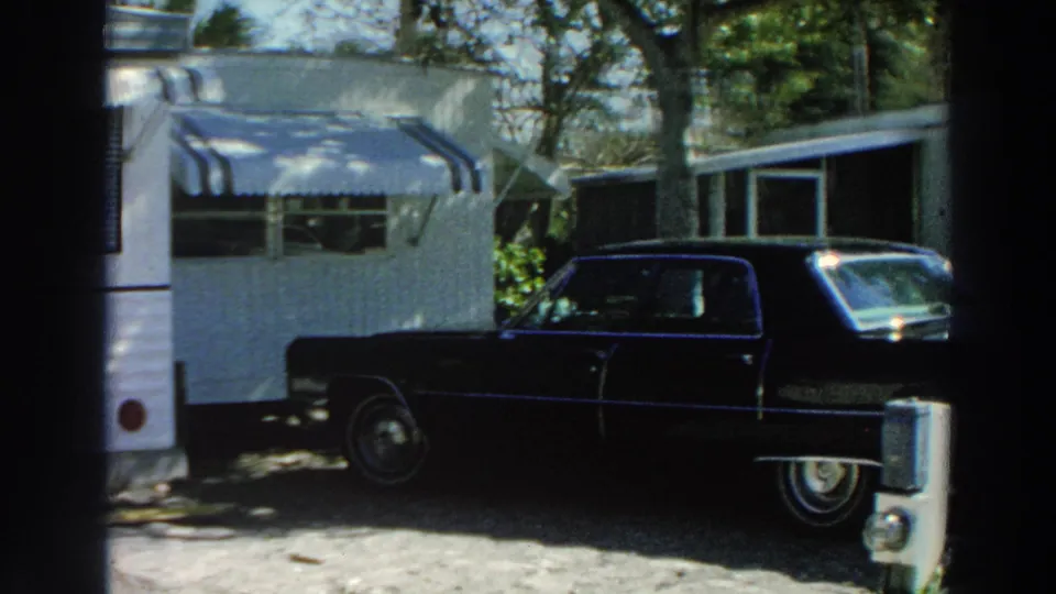 1966 Puerto Rico home-movie frame with a dark Cadillac-style sedan parked next to a white mobile home and trees.