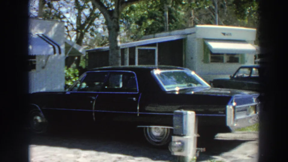 Final 8mm frame of a dark Cadillac-style sedan, white mobile homes, awnings, trees, and sunlit chrome in Puerto Rico.