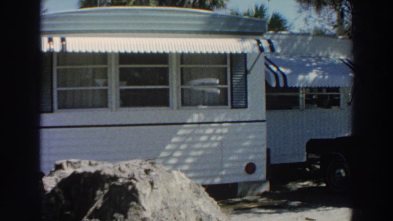 1966 Puerto Rico home-movie frame of a white mobile home with striped awnings, trees, and earth piled in the foreground.