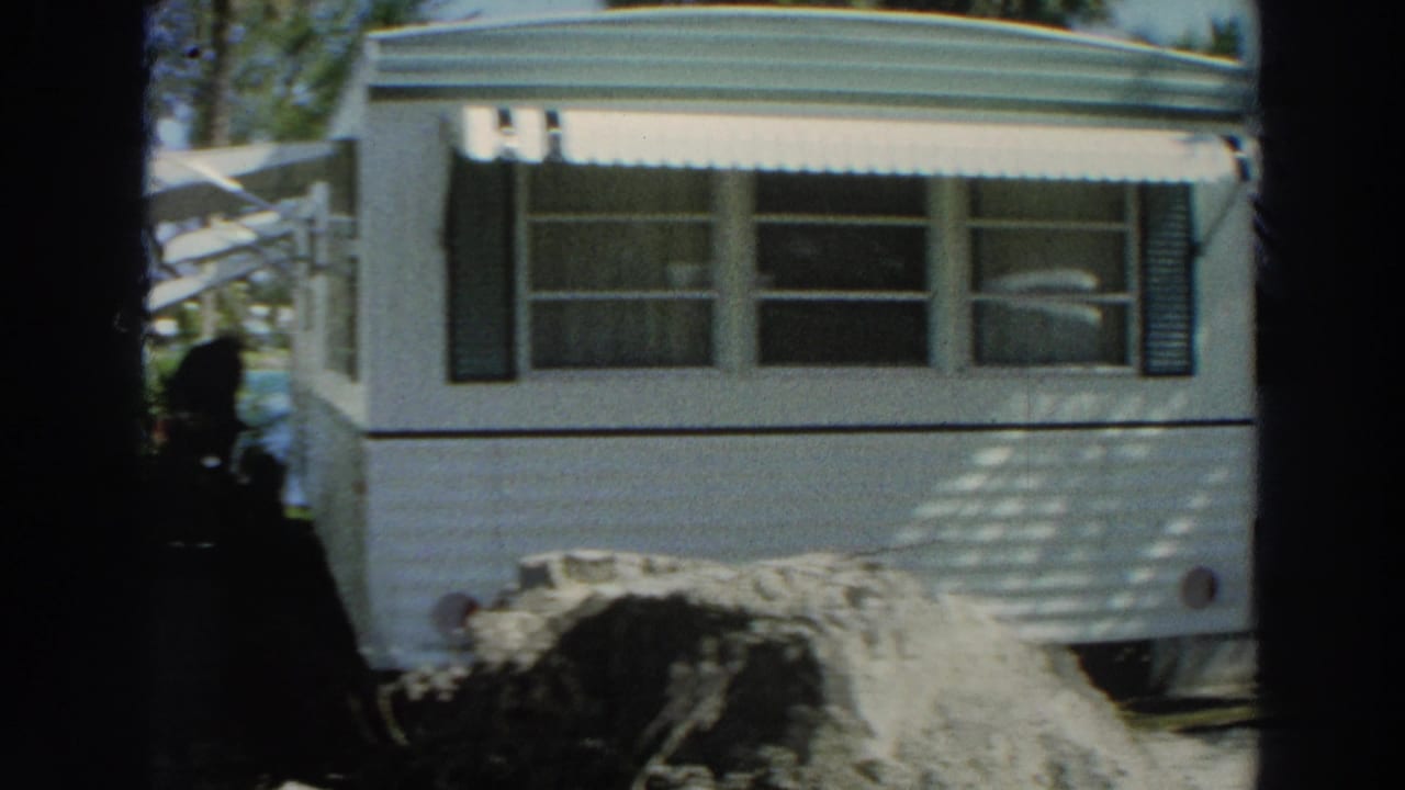 1966 Puerto Rico frame showing the mobile home facade, striped awning, foreground earth, and a dark figure near the left edge.