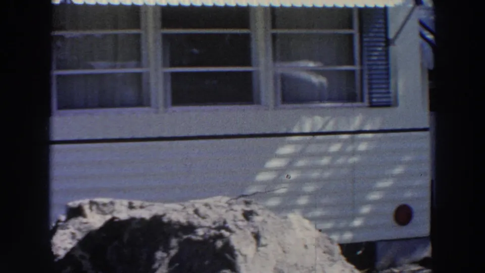 1966 Puerto Rico home-movie frame showing the side of a white mobile home with windows, shutters, and mottled sunlight.