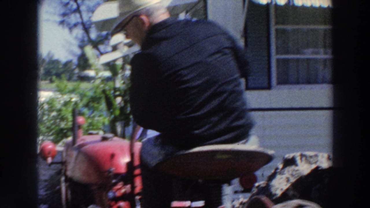 In 1966 Puerto Rico footage, an adult man drives a red tractor past a white mobile home in bright daylight.