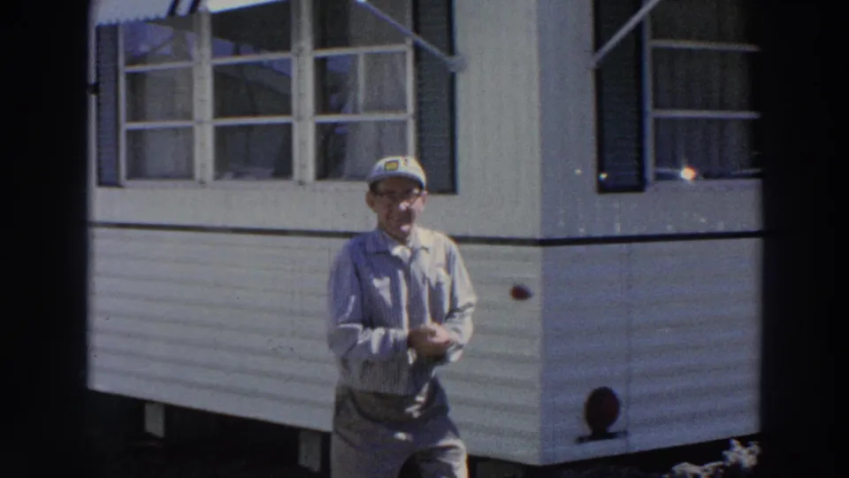 The man stands facing the camera in front of the white mobile home, with windows and shutters behind him.