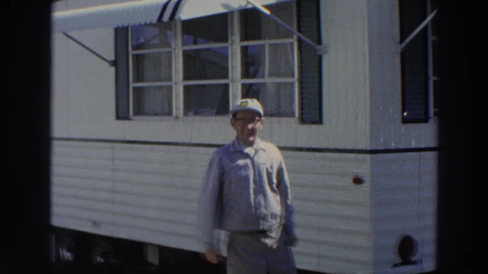 The man appears to smile or speak toward the camera while standing outside the white mobile home in Puerto Rico.