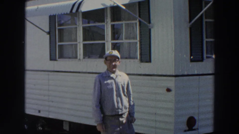 In Puerto Rico, 1966, an adult man in a white cap and glasses stands near a white mobile home with shutters and an awning.