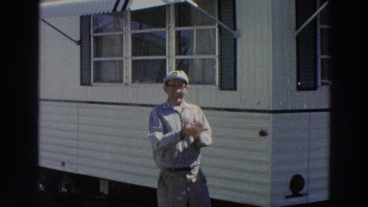 In Puerto Rico, 1966, the man in a cap and glasses rubs his hands in front of a white mobile home exterior.