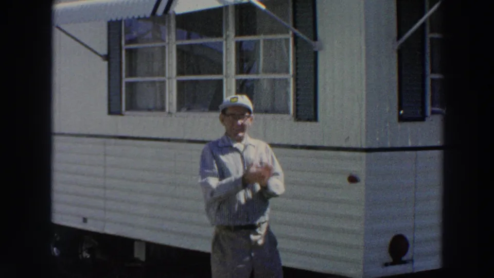In Puerto Rico, 1966, the man in a cap and glasses rubs his hands in front of a white mobile home exterior.