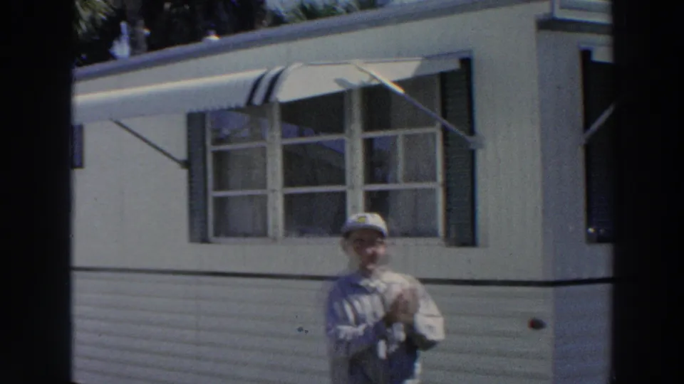 In Puerto Rico, 1966, the man remains outside the white mobile home, partly low in frame, with windows and an awning behind him.