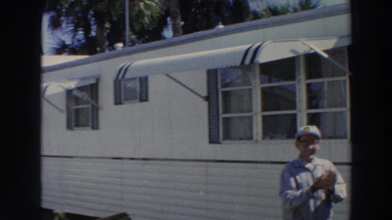 Smiling man standing beside a white mobile home with striped awnings and palm trees behind it.