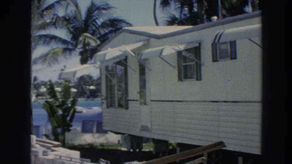 A 1966 Puerto Rico home-movie view shows a white mobile home with open windows, striped awnings, palm trees, and blue water beyond it.