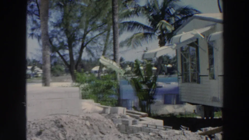 A Puerto Rico 1966 home-movie frame shows the side of a white mobile home near sandy ground, palms, construction blocks, and blue water.