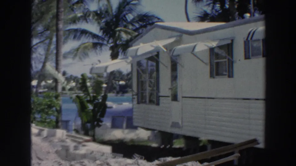1966 Puerto Rico 8mm frame showing a tan mobile home with awnings, windows, palms, and tropical foliage.
