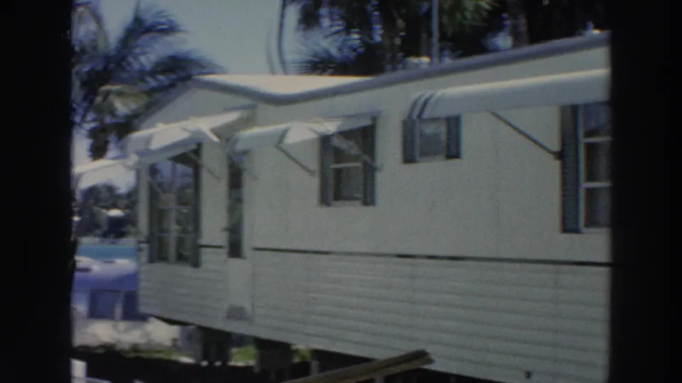 1966 Puerto Rico home-movie frame with the side of a tan mobile home, shaded windows, awnings, and palm trees.