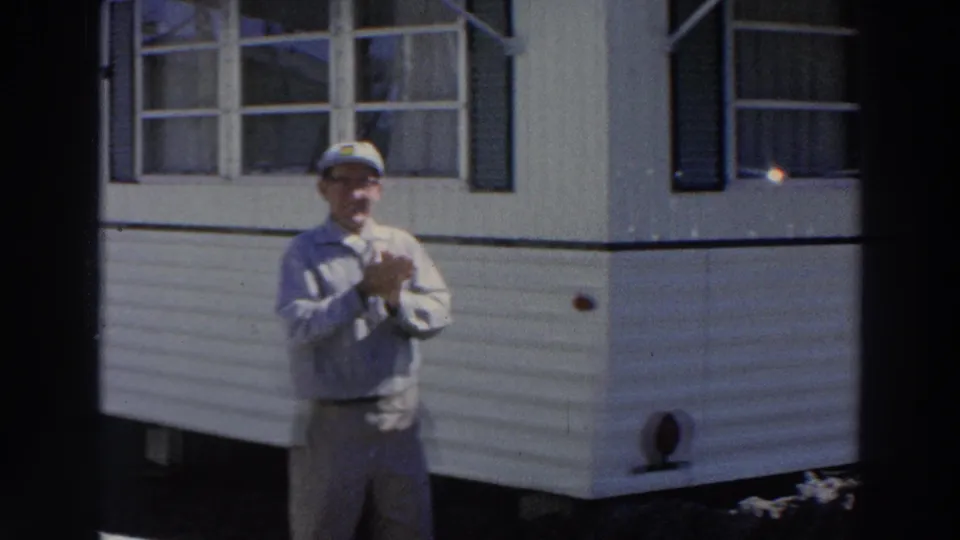 1966 Puerto Rico 8mm frame of an adult man in cap and light shirt beside a tan mobile home, hands held near his chest.