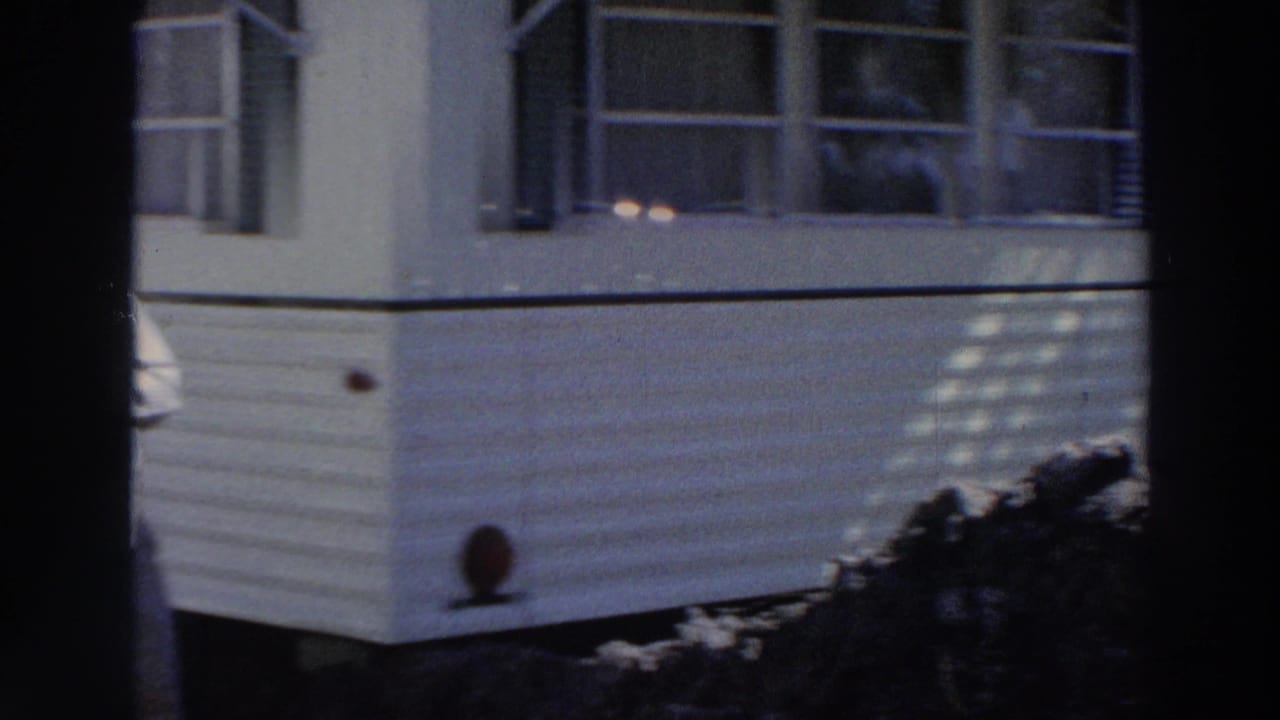 1966 Puerto Rico 8mm frame showing a tan mobile home exterior with white siding, dark shutters, shrubs, and partial awning shade.