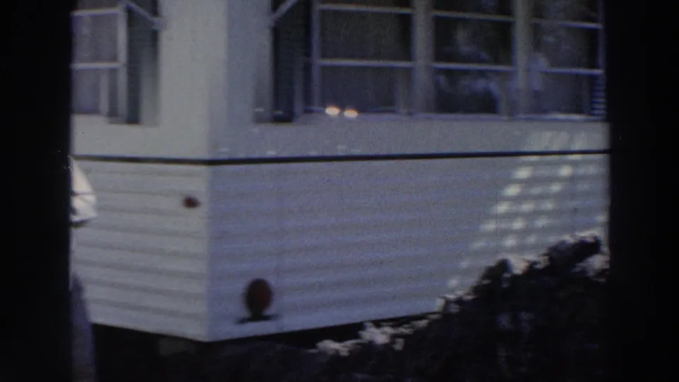 1966 Puerto Rico 8mm frame showing a tan mobile home exterior with white siding, dark shutters, shrubs, and partial awning shade.