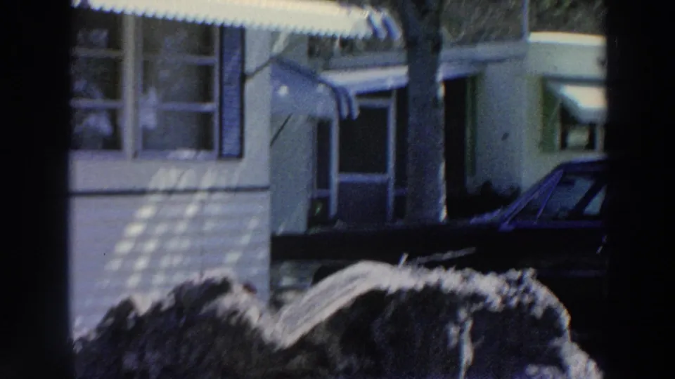 1966 Puerto Rico home-movie frame with a mobile home entrance, striped awning, nearby shrubs, tree trunk, and dark parked car.