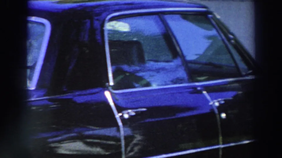 Blue Cadillac parked outside a light-colored Puerto Rico building, seen in a grainy 1966 8mm home-movie frame.