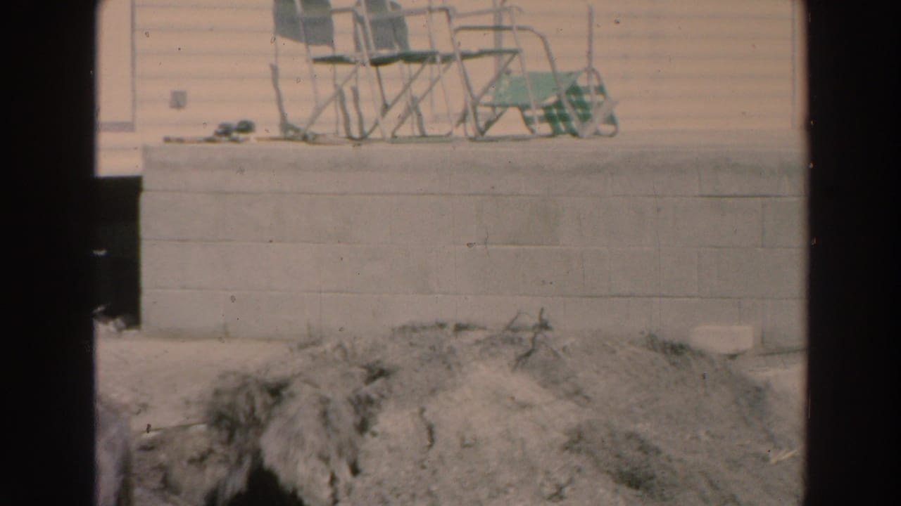 1966 Puerto Rico yard view with a concrete block wall, loose dirt, and stacked metal lawn chairs near a house exterior.