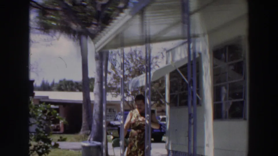 1966 Puerto Rico home-movie frame showing an adult woman in a patterned dress near a covered porch and white building.