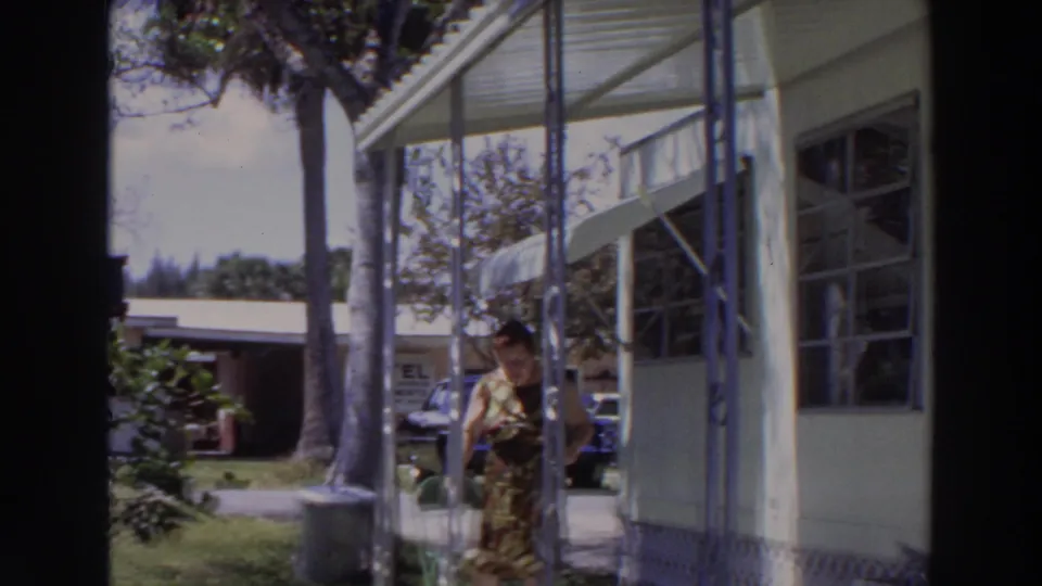 1966 Puerto Rico frame of an adult woman beside a screened porch, with palm trees and parked cars behind her.