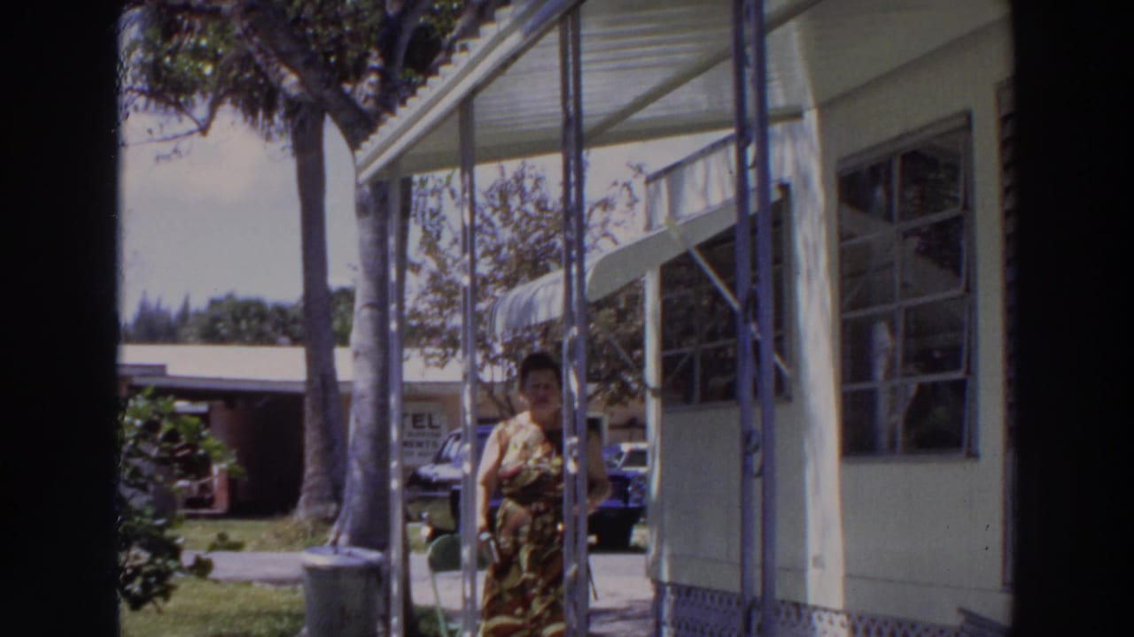 1966 Puerto Rico home-movie frame showing a woman on a porch near metal supports, palm trees, and a parked car.
