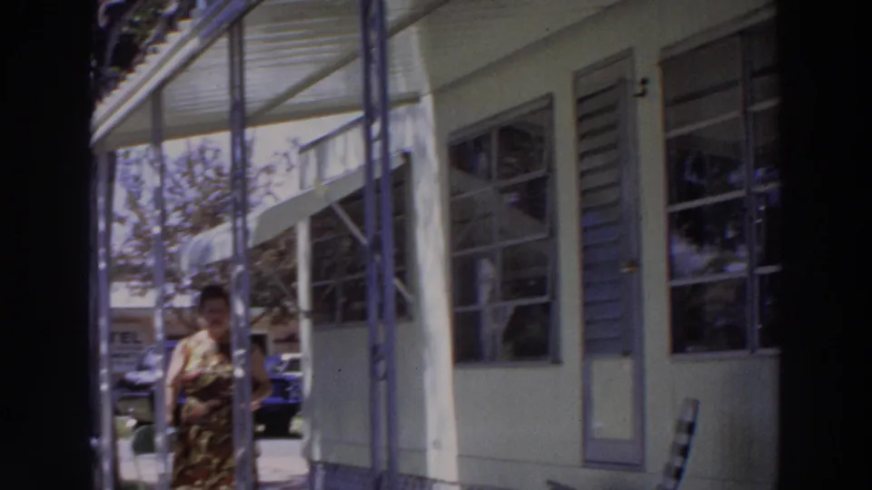1966 Puerto Rico frame with a woman near a porch column, louvered windows, a door, and a parked car in the background.