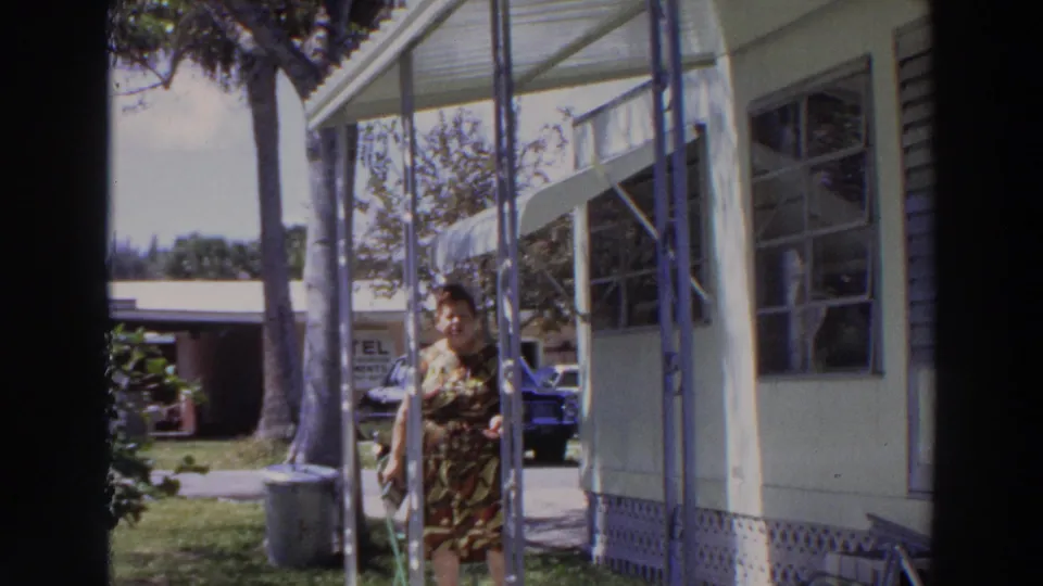 In 1966 Puerto Rico footage, a woman in a patterned dress walks near a covered mobile home porch with palm trees and parked cars behind her.