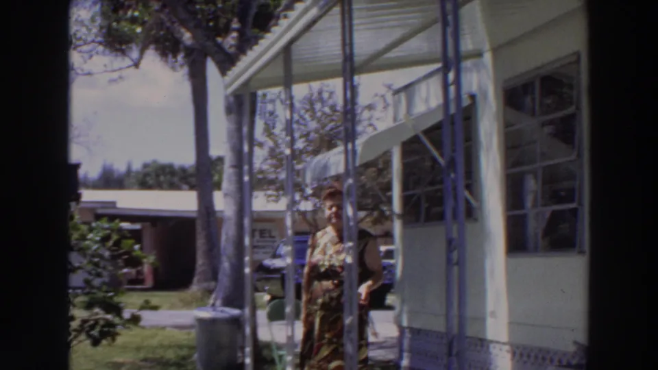 The woman is partly obscured by a porch post while walking beside the mobile home, with a yard, palms, and distant signage behind her.