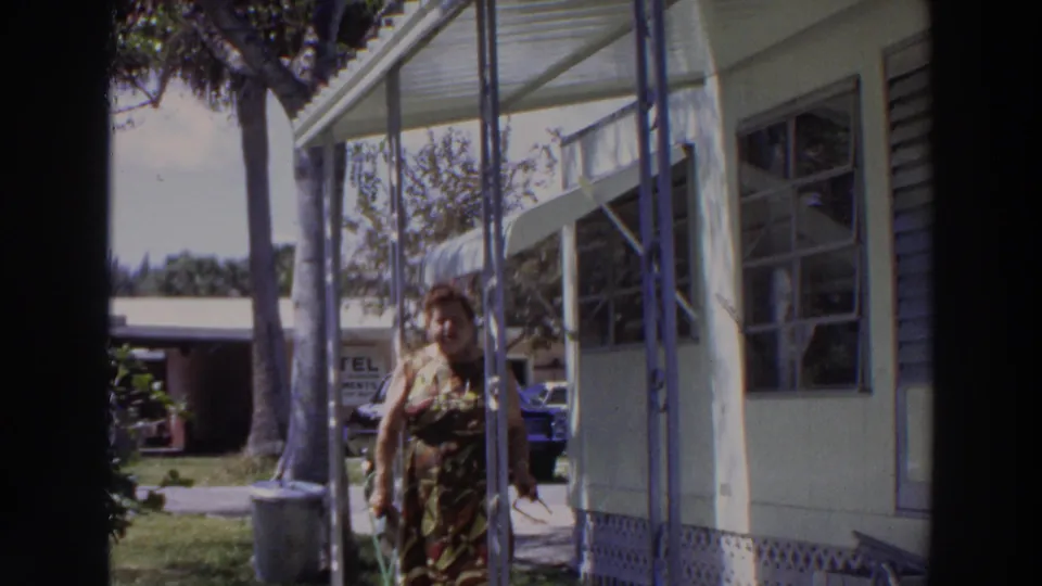 Near the end of the clip, the woman walks past the porch of a light mobile home in 1966 Puerto Rico, with palms, grass, and parked cars visible.