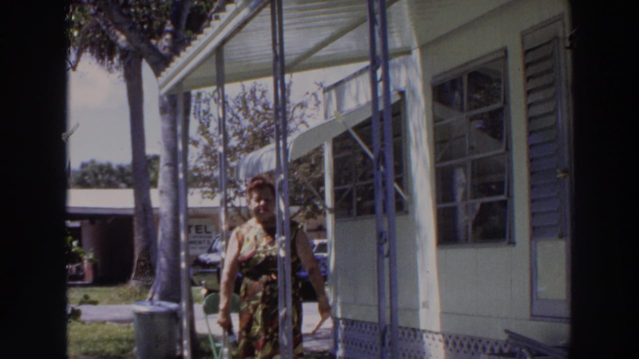 In 1966 Puerto Rico footage, a woman in a patterned dress walks toward the camera on a covered porch with palms and a car behind her.