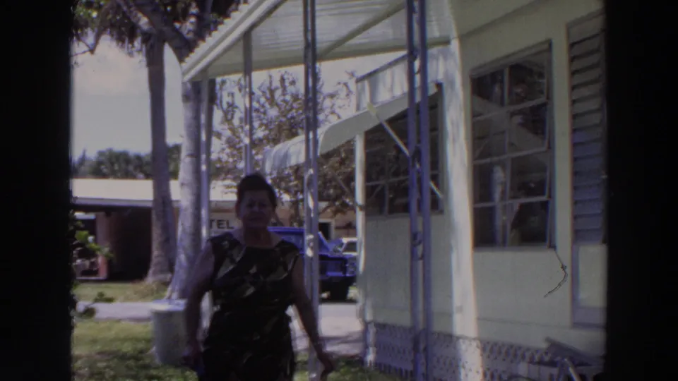 The woman nears the camera on the porch, partly in shadow, framed by the house exterior, windows, palm trunks, lawn, and parked car.