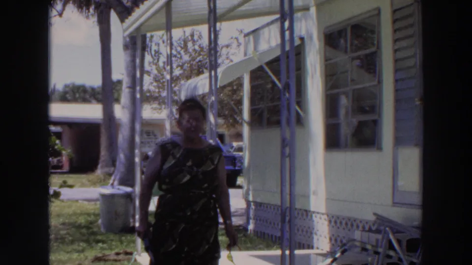 Near the end of the shot, the woman stands close to the camera on the porch, with a light house exterior, palms, yard, and parked car behind her.