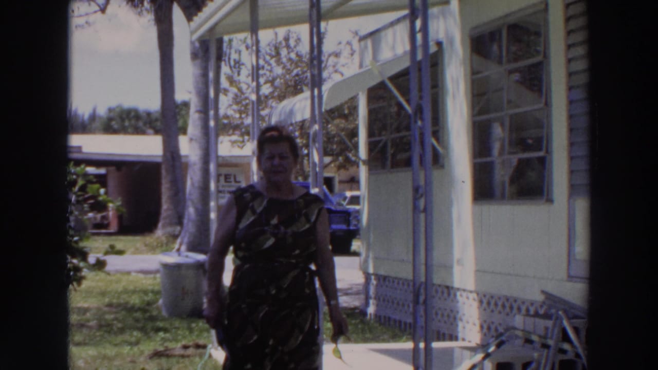 Woman in a sleeveless patterned dress walking on a porch beside a light-colored mobile home.