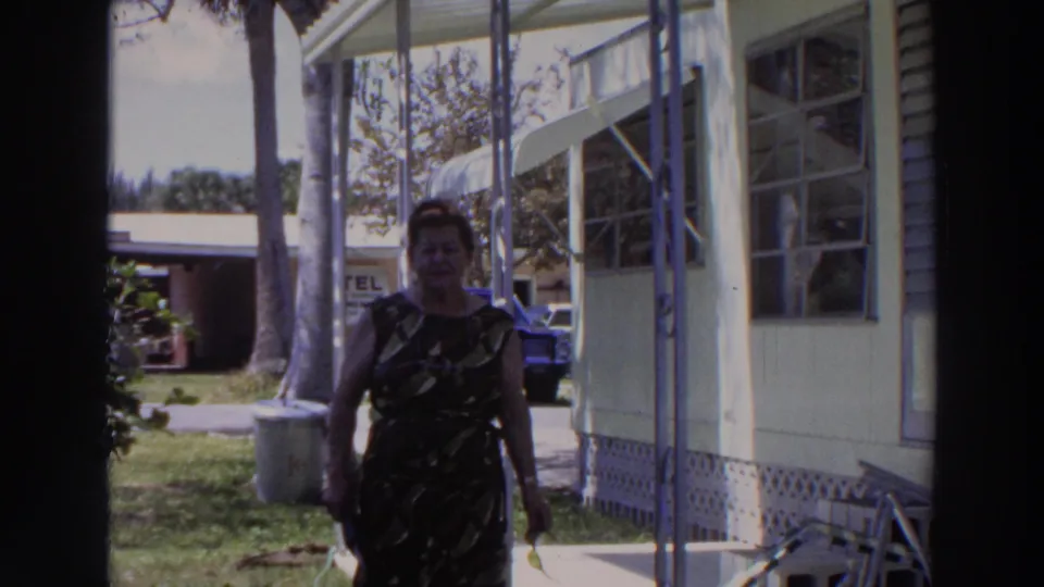 In 1966 Puerto Rico, a woman walks toward the camera beside a pale mobile home with palm trees, lawn, and a partial TEL sign behind her.