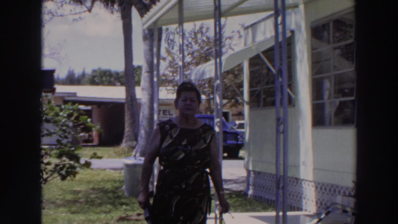 Woman walking closer to the camera beside porch posts and windows of a mobile home.