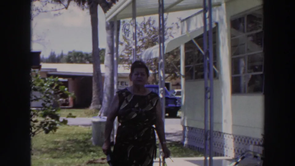 The woman continues toward the camera on the mobile-home porch, with the lawn, palm trunks, blue vehicle, and partial background sign visible.