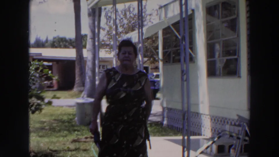 The woman is closer in frame, walking along the sunlit porch area of a Puerto Rico mobile home with palms and lodging signage behind her.